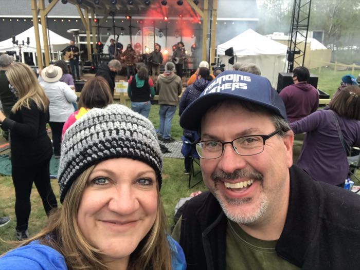 Trevor and Kat at an outdoor festival, with The Infamous Stringdusters playing on the stage behind them.